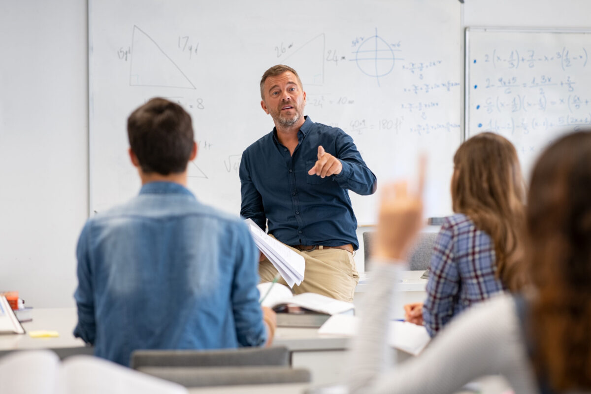 stock-photo-professor-pointing-at-college-student-with-hand-raised-in-classroom-student-raising-a-hand-with-a-1836272266